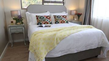 Cama bem arrumada com manta amarela estampada, dois travesseiros decorativos, abajur e mesinha de cabeceira com um vaso de flores.