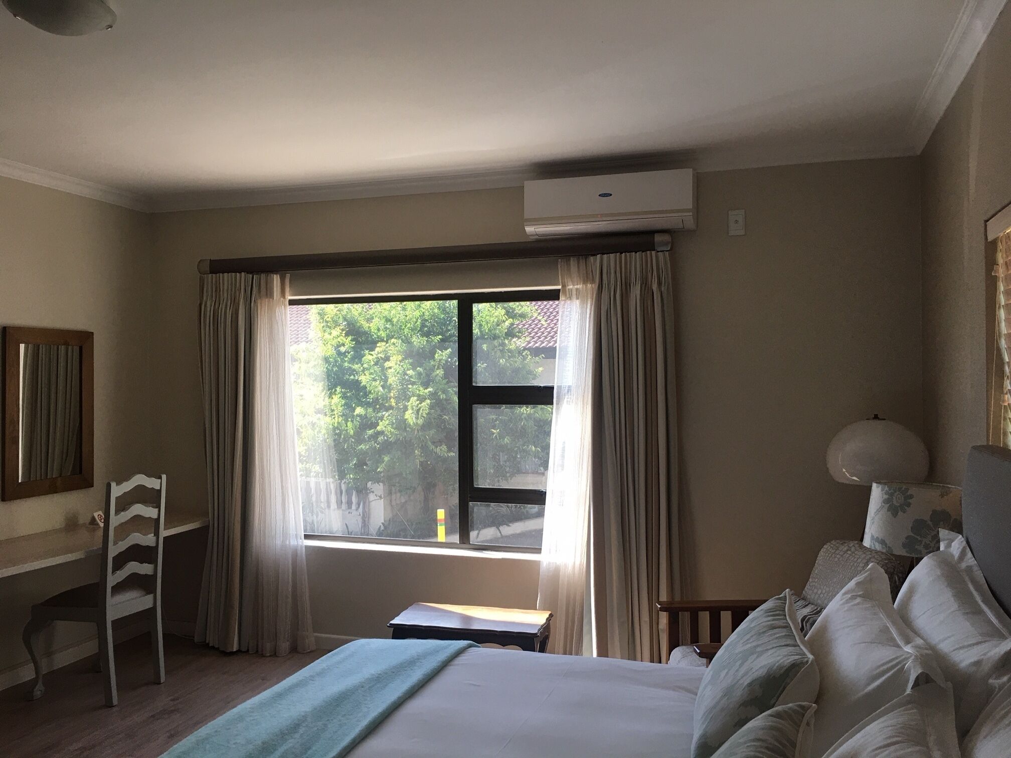A hotel room with a bed, a desk, a chair, a window with curtains, and an air conditioning unit.