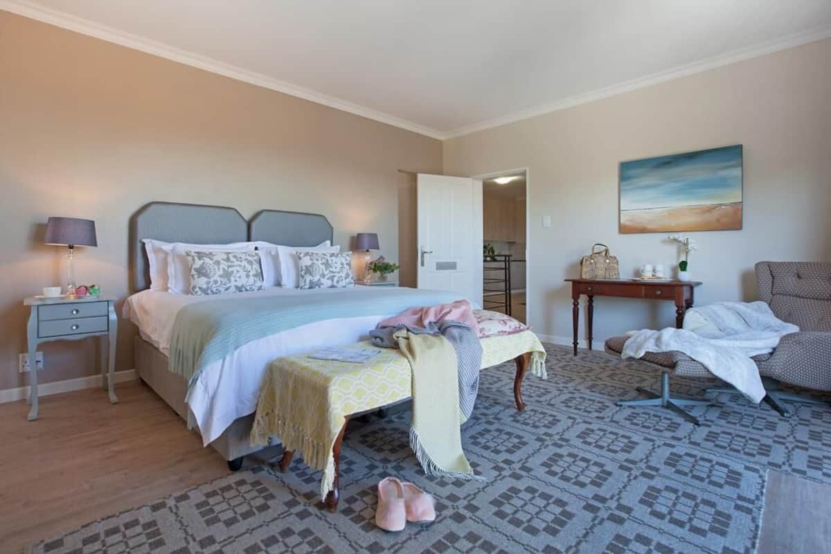 Quarto com cama, mesas de cabeceira, um banco, uma cadeira, escrivaninha e um quadro na parede.