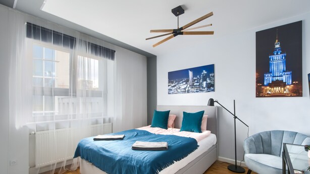Une chambre à coucher moderne avec un lit, une lampe sur pied, une chaise et un tableau représentant un paysage urbain.