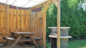 Une table de pique-nique et un barbecue en bois sont installés dans un jardin, avec une clôture en bois et de la verdure en arrière-plan.