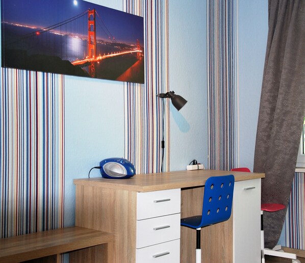 Un dormitorio con un escritorio de madera, una silla azul, una cama con cabecera gris y una pared a rayas.