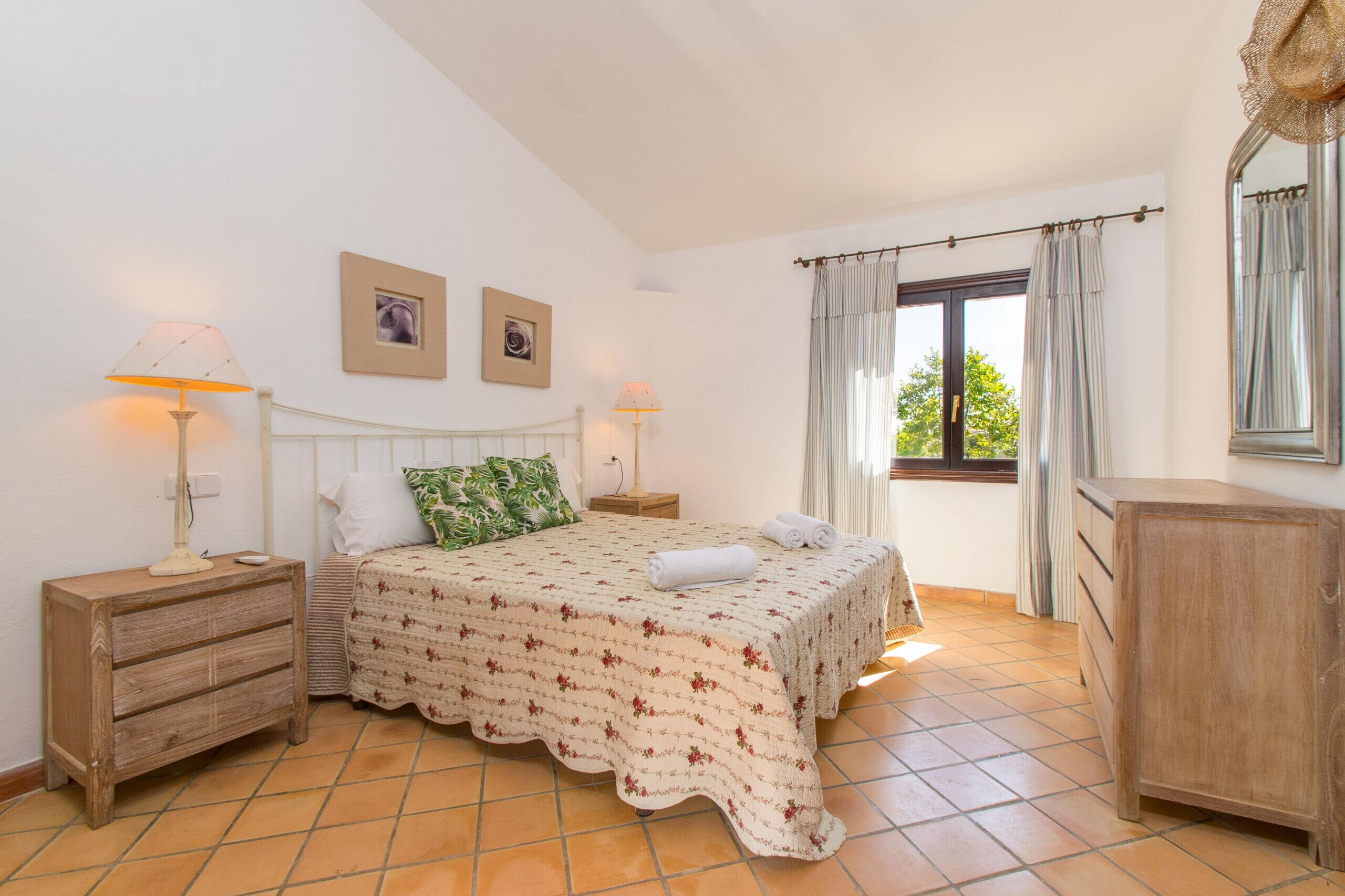 Une chambre à coucher comprenant un lit, deux tables de chevet, une commode, une coiffeuse, une fenêtre avec des rideaux et un miroir fixé au mur.