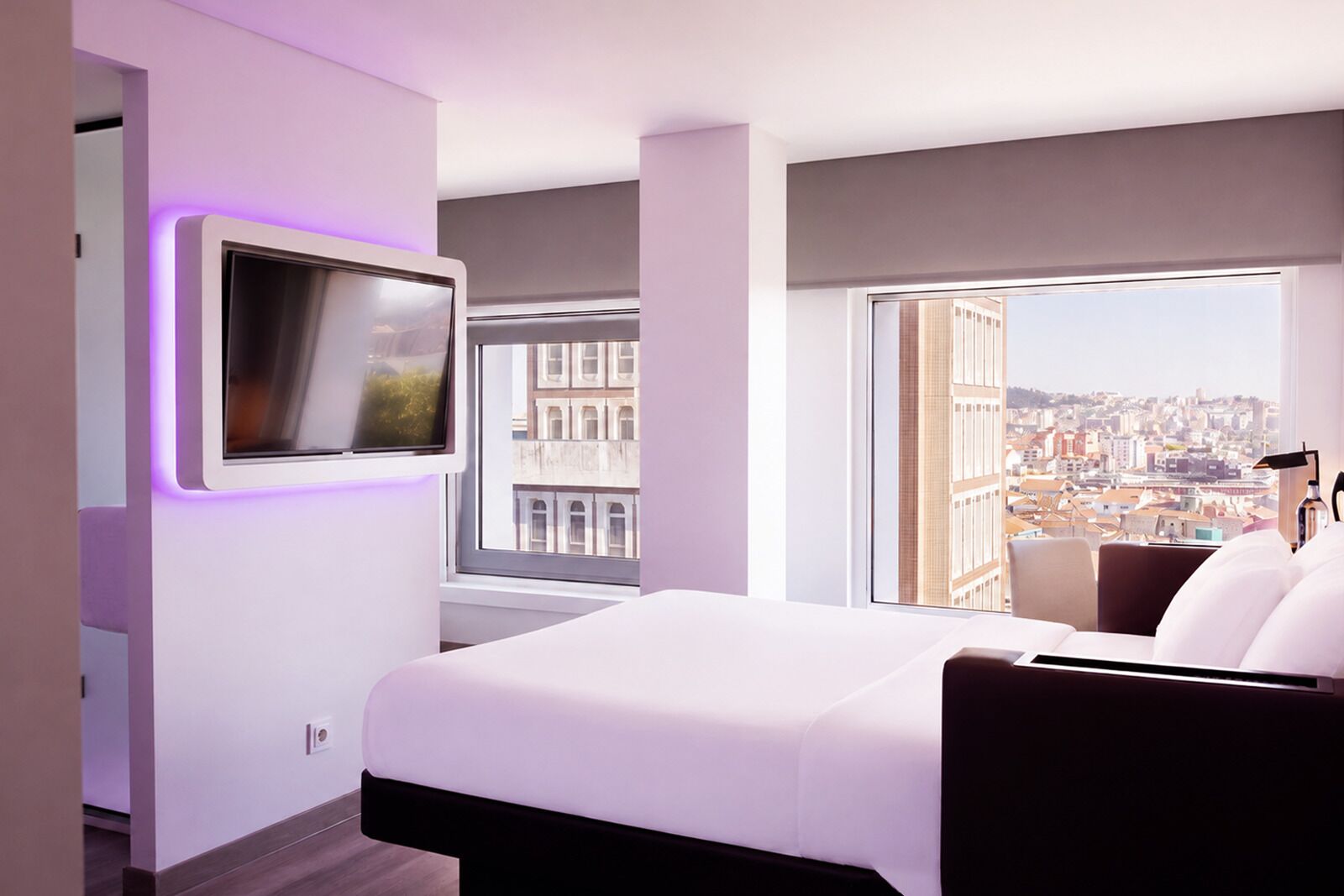 A modern hotel room with a large bed, a flat-screen TV mounted on the wall, and a city view through the window.