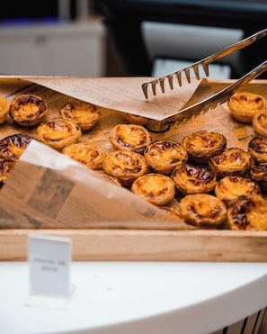 A tray of freshly baked pastries, likely Portuguese egg tarts, with a pair of tongs resting on top.