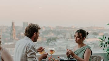 Una mesa de comedor con dos personas sentadas, un paisaje urbano al fondo y un cielo despejado.