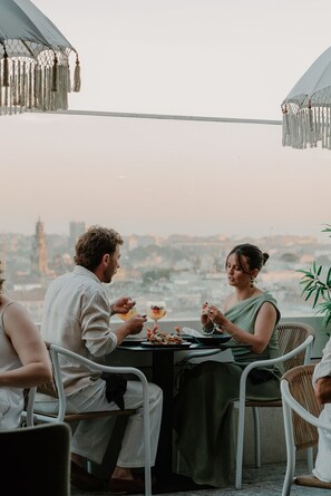 A dining table with two people seated, a cityscape in the background, and a clear sky. - YOTEL Porto (Porto)