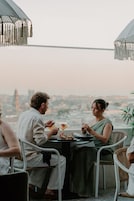 Una mesa de comedor con dos personas sentadas, un paisaje urbano al fondo y un cielo despejado.