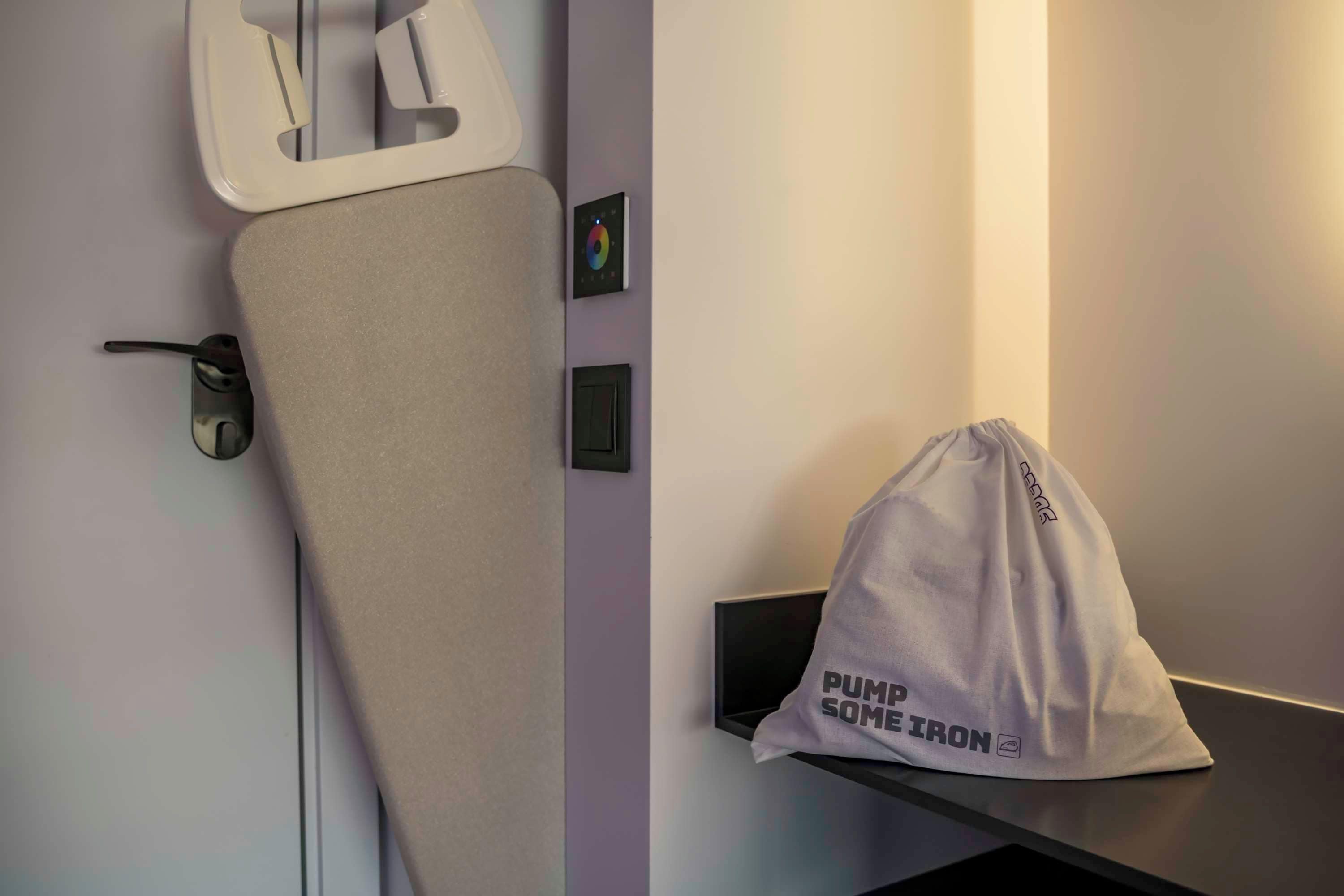 Una habitación con una cama, una mesita de noche, un panel de control en la pared y una bolsa con el texto 'PUMP SOME IRON' sobre la mesa.