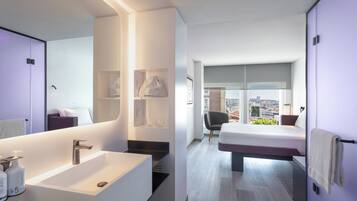 A modern hotel room with a large bathroom, a bed with a city view, and a small seating area.