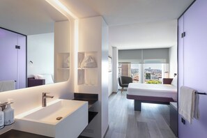 A modern hotel room with a large bathroom, a bed with a city view, and a small seating area.