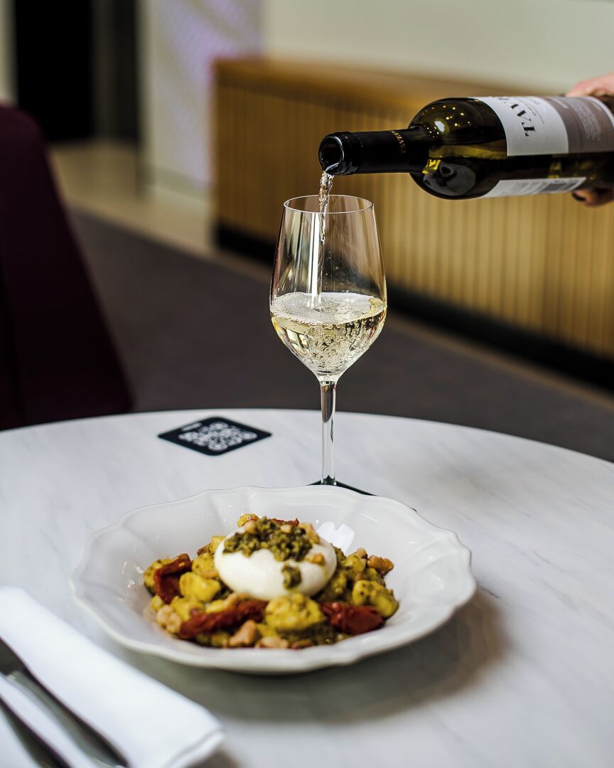 Une bouteille de vin blanc est versée dans un verre posé sur une table, à côté d’une assiette de nourriture.