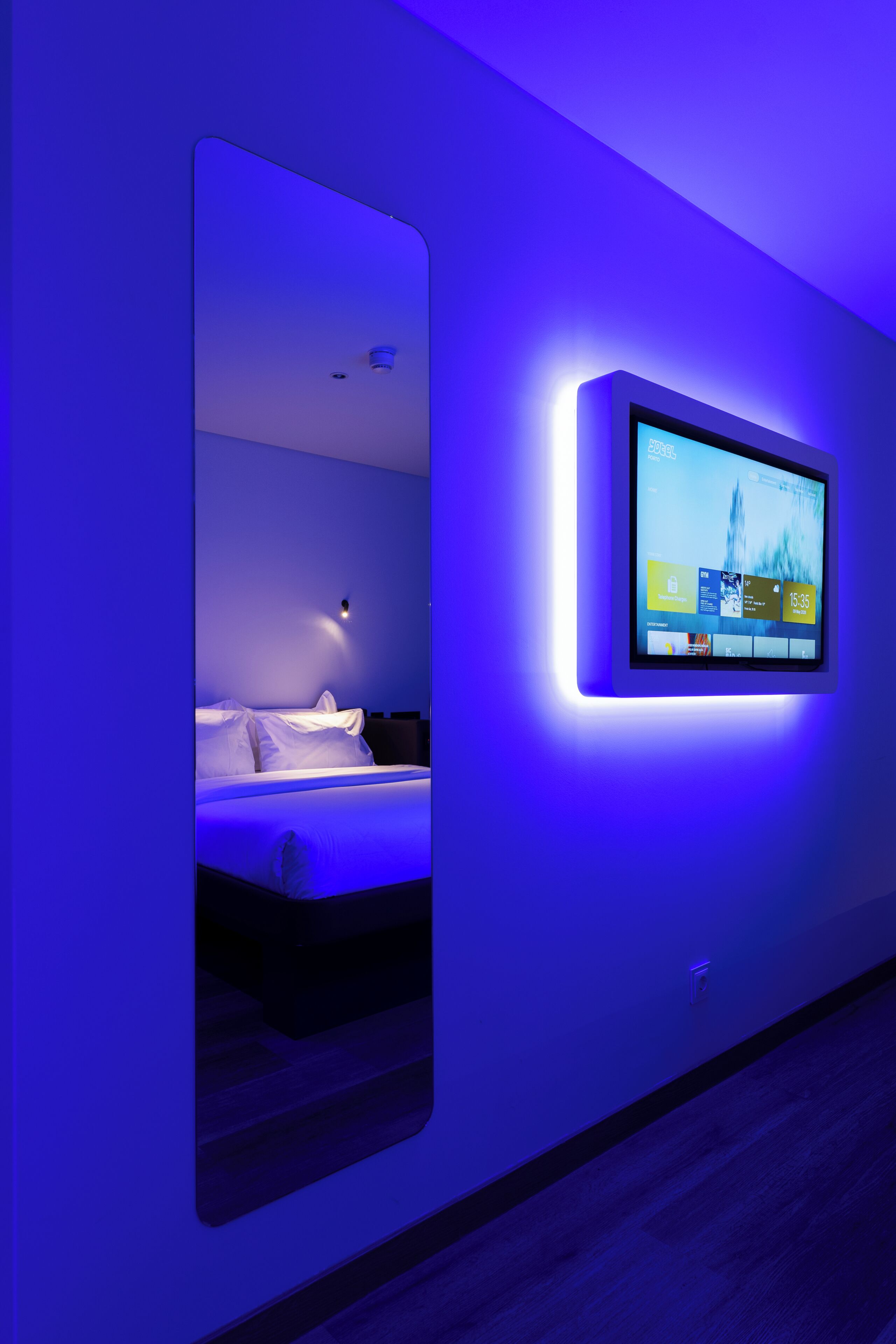 A modern hotel room with a large mirror, a flat-screen TV mounted on the wall, and a bed with white linens.