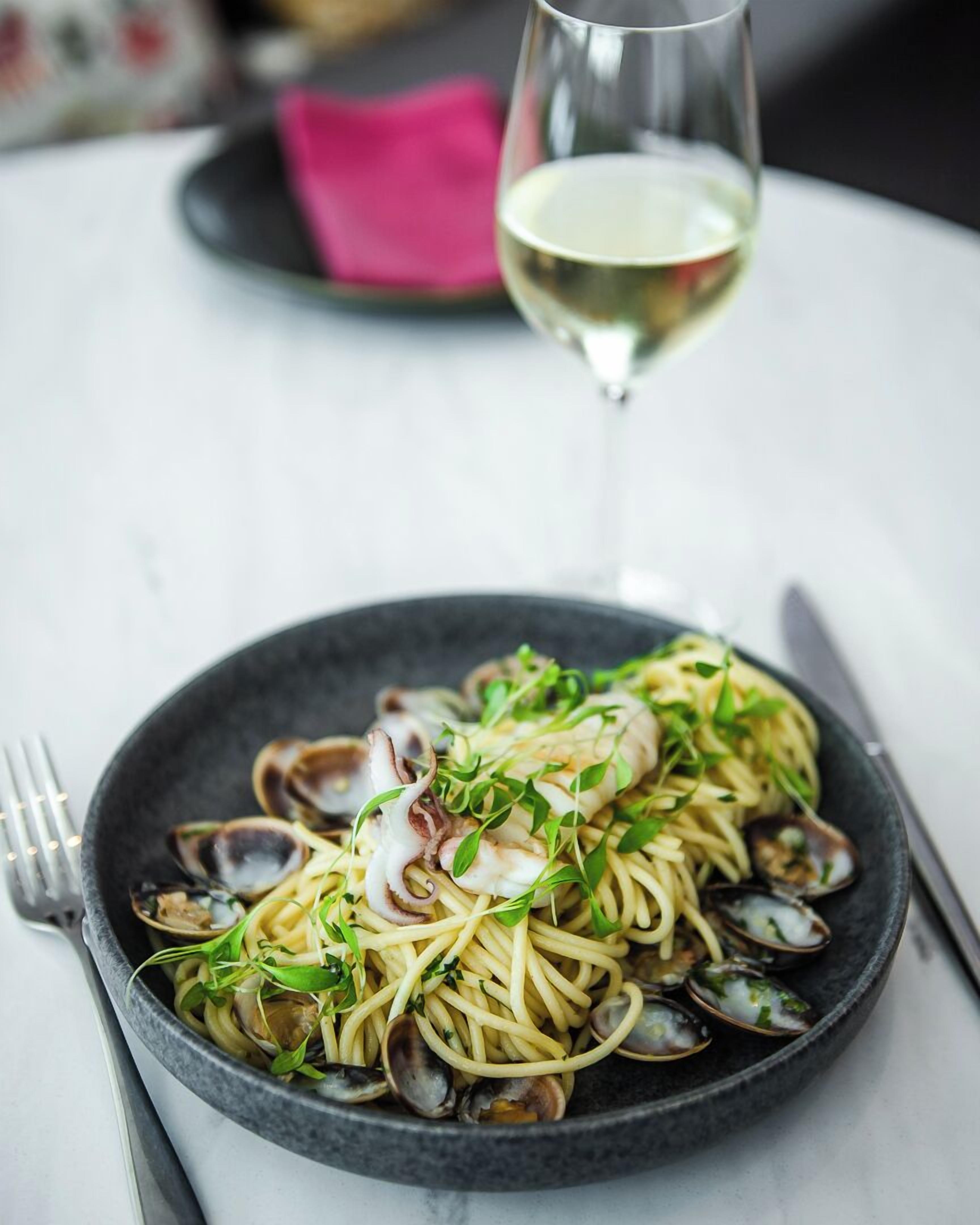 A plate of pasta with clams and greens, a glass of white wine, and a fork on a table.