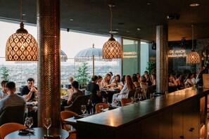 A restaurant with a long counter, hanging patterned lamps, and a view of the cityscape. - YOTEL Porto (Porto)