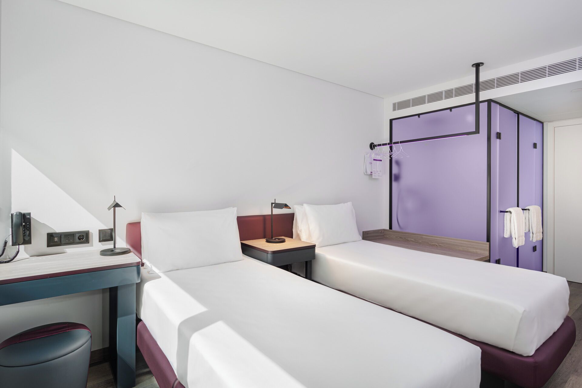 Une chambre d’hôtel avec deux lits simples, un bureau avec une lampe, un téléphone et une petite poubelle.
