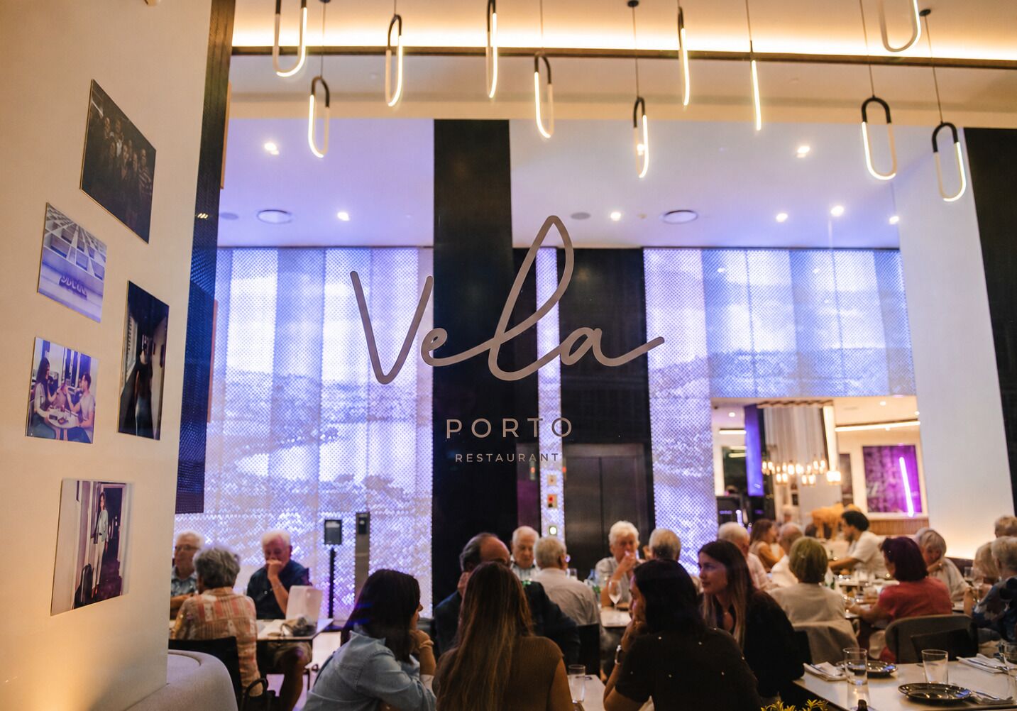 An indoor dining area with tables and patrons, a large illuminated sign reading 'Vela PORTO RESTAURANT', and a modern interior design with hanging lights.