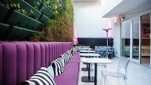 A modern outdoor seating area with purple cushioned benches and clear plastic chairs, surrounded by greenery and a glass door leading to an interior space. - YOTEL Porto (Porto)