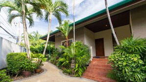A pathway leading to a building with a green roof and a brick wall. There are various plants and a statue along the path. - Viola Pool Villa (Rawai)