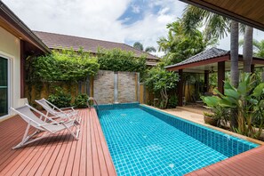 A swimming pool with a wooden deck and lounge chairs, surrounded by greenery and a stone wall.