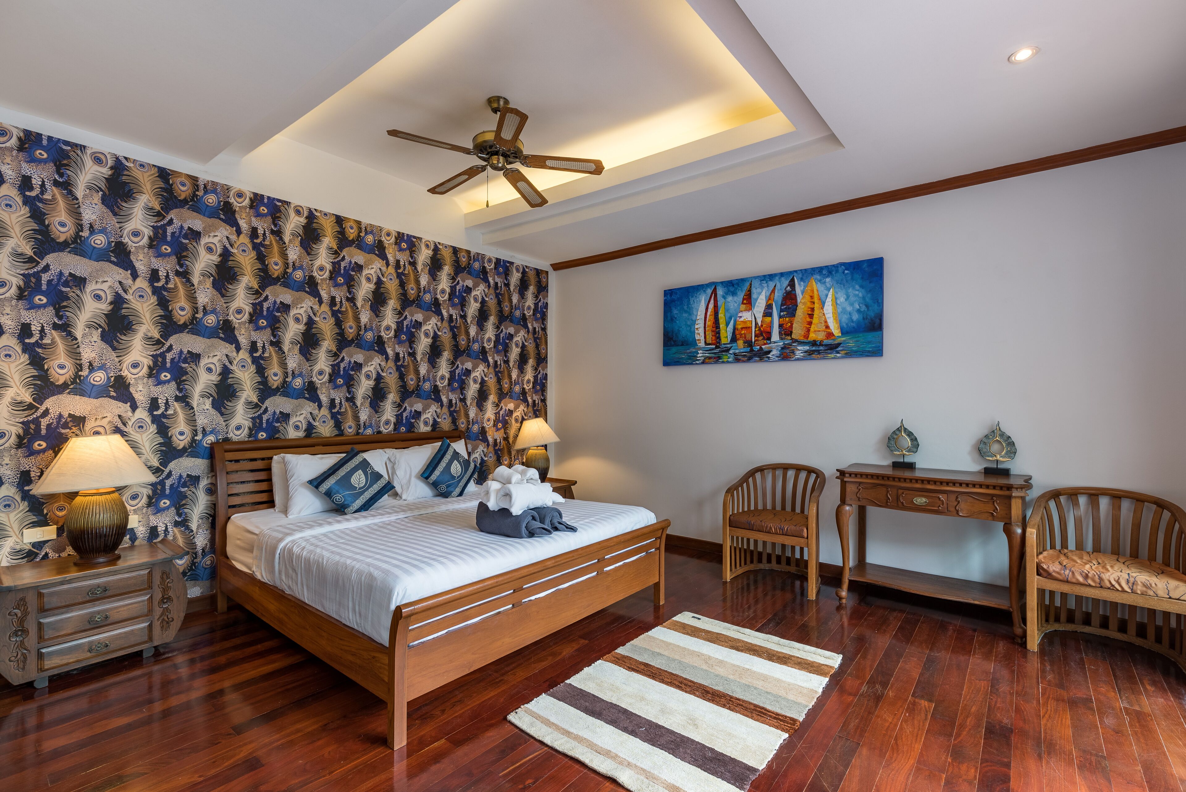 A bedroom with a wooden bed, a nightstand with a lamp, a desk with two chairs, a painting of sailboats, and a patterned wallpaper.