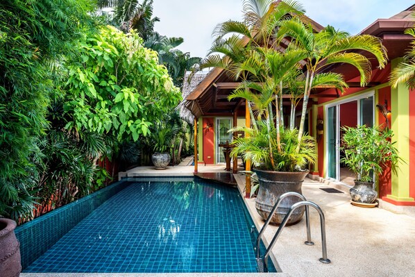 A swimming pool surrounded by lush greenery and potted plants, with a building featuring a red and yellow facade in the background. - Chaba Pool Villa (Rawai)