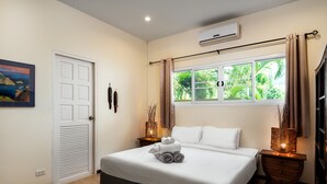 Una cama bien tendida con toallas enrolladas, una ventana con cortinas, una mesita de noche con lámpara y un aire acondicionado fijado a la pared.