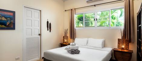 Una cama bien tendida con toallas enrolladas, una ventana con cortinas, una mesita de noche con lámpara y un aire acondicionado fijado a la pared.