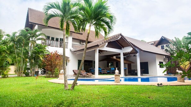 Una casa de dos pisos con piscina, rodeada de exuberante vegetación y palmeras.