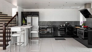 A modern kitchen with black cabinetry, a white island, stainless steel appliances, and a staircase with wooden railings. - Sky Pool Villa (Rawai)