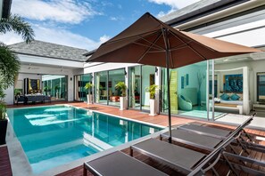 Uma área moderna com piscina ao ar livre, cadeiras de descanso e um guarda-sol grande, cercada por portas de vidro e plantas em vasos.