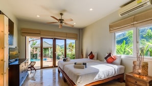 Ein Schlafzimmer mit einem großen Bett, einem Deckenventilator, einer Klimaanlage, einem Nachttisch aus Holz und Blick auf einen Swimmingpool und Grünfläche.