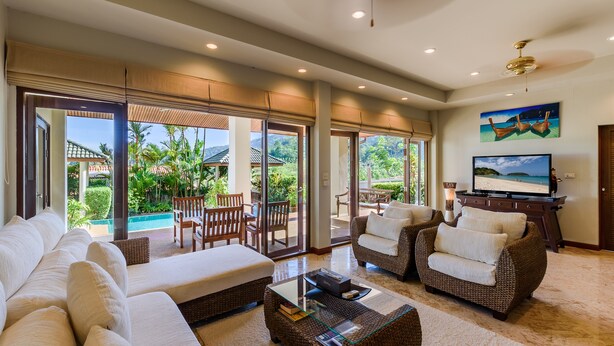 Ein großzügiger Wohnbereich mit einer großen Couch, zwei Sesseln, einem Couchtisch und einem Fernseher. Große Fenster bieten Blick auf einen Pool und Grünflächen.