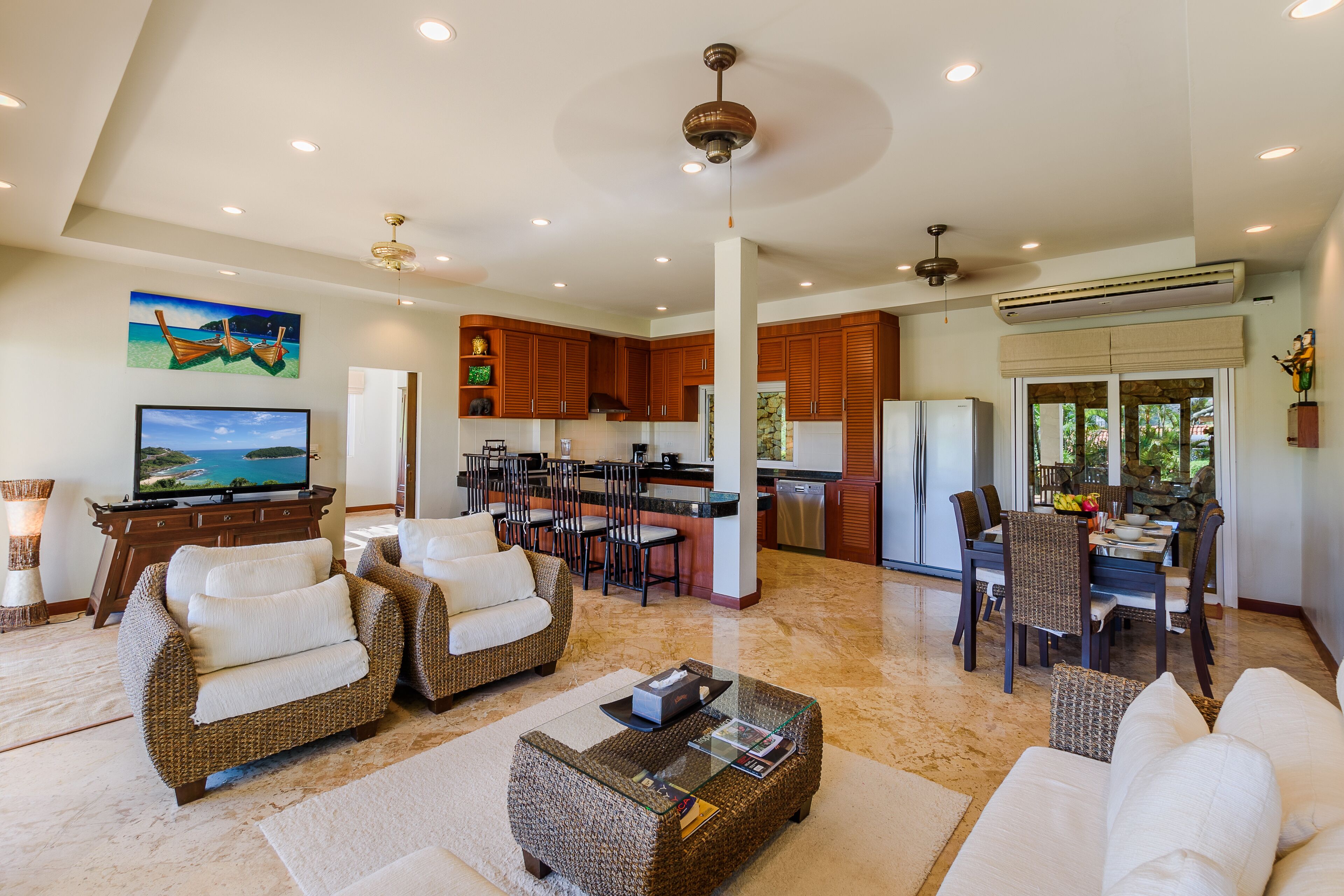 A spacious living area with a large flat-screen TV, wicker furniture, and a kitchen in the background.