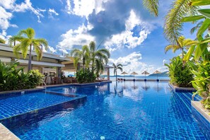 A swimming pool with clear blue water, surrounded by palm trees and lounge chairs, with a view of the ocean and mountains in the background. - Serenity 2 bedroom Condominium (Rawai)