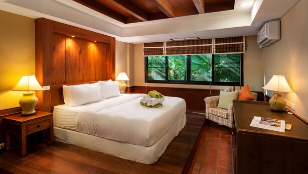 A hotel room with a large bed, two bedside tables with lamps, a wooden headboard, a window with a view of greenery, and a desk with a lamp. - The Sands 2 bedroom apartment (Rawai)