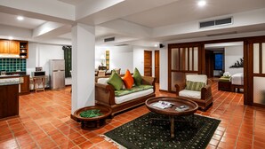 A spacious living area with a sofa, armchairs, and a coffee table. A kitchen with wooden cabinets and a microwave is visible in the background. - The Sands 2 bedroom apartment (Rawai)