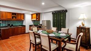 A dining area with a table set for four, surrounded by wooden chairs. A television is mounted on the wall, and a lamp is on a side table. - The Sands 2 bedroom apartment (Rawai)