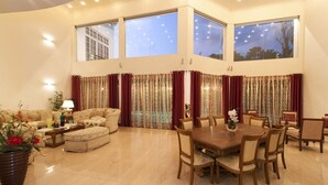A spacious living room with a large dining table, multiple sofas, and a coffee table. There are large windows with curtains, a potted plant, and a lamp on a side table. - Villa 53 (Battaramulla)