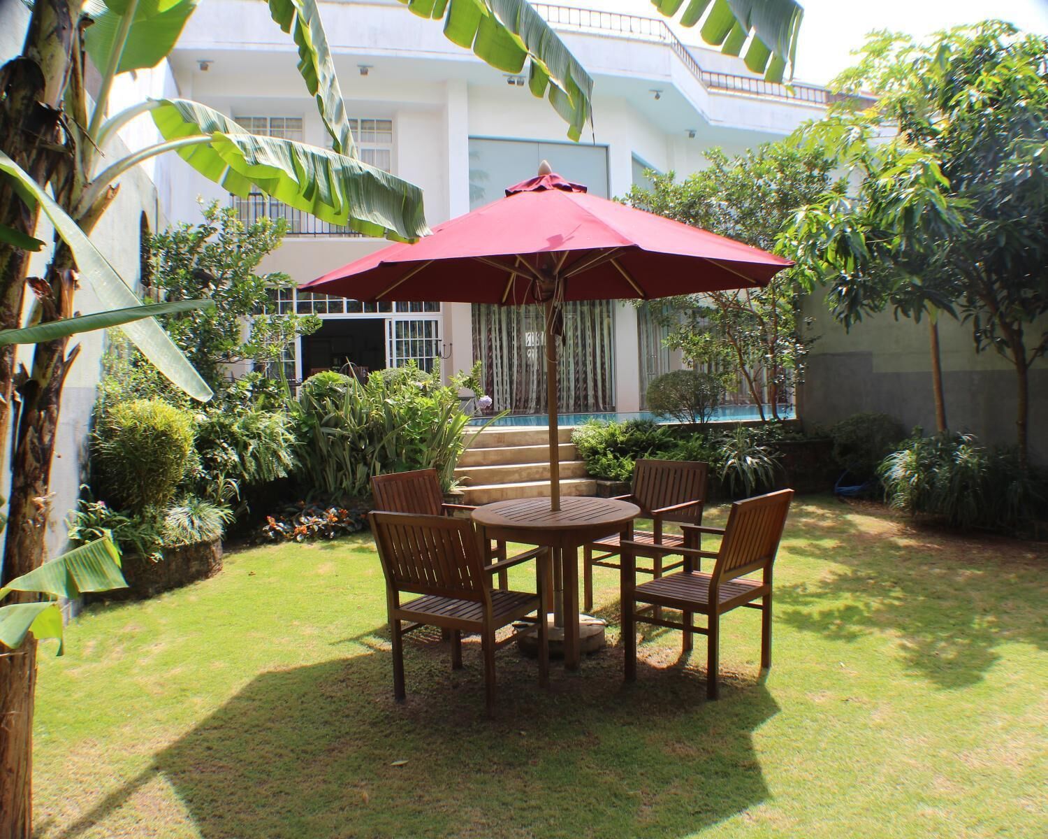 A patio with a round wooden table and chairs, a red umbrella, and a view of a building with steps and a pool.