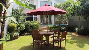 A patio with a round wooden table and chairs, a red umbrella, and a view of a building with steps and a pool.