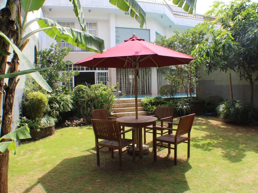 A patio with a round wooden table and chairs, a red umbrella, and a view of a building with steps and a pool.