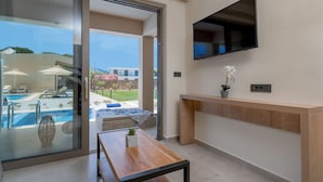 A modern living room with a TV, a wooden table, and a view of a swimming pool and garden.