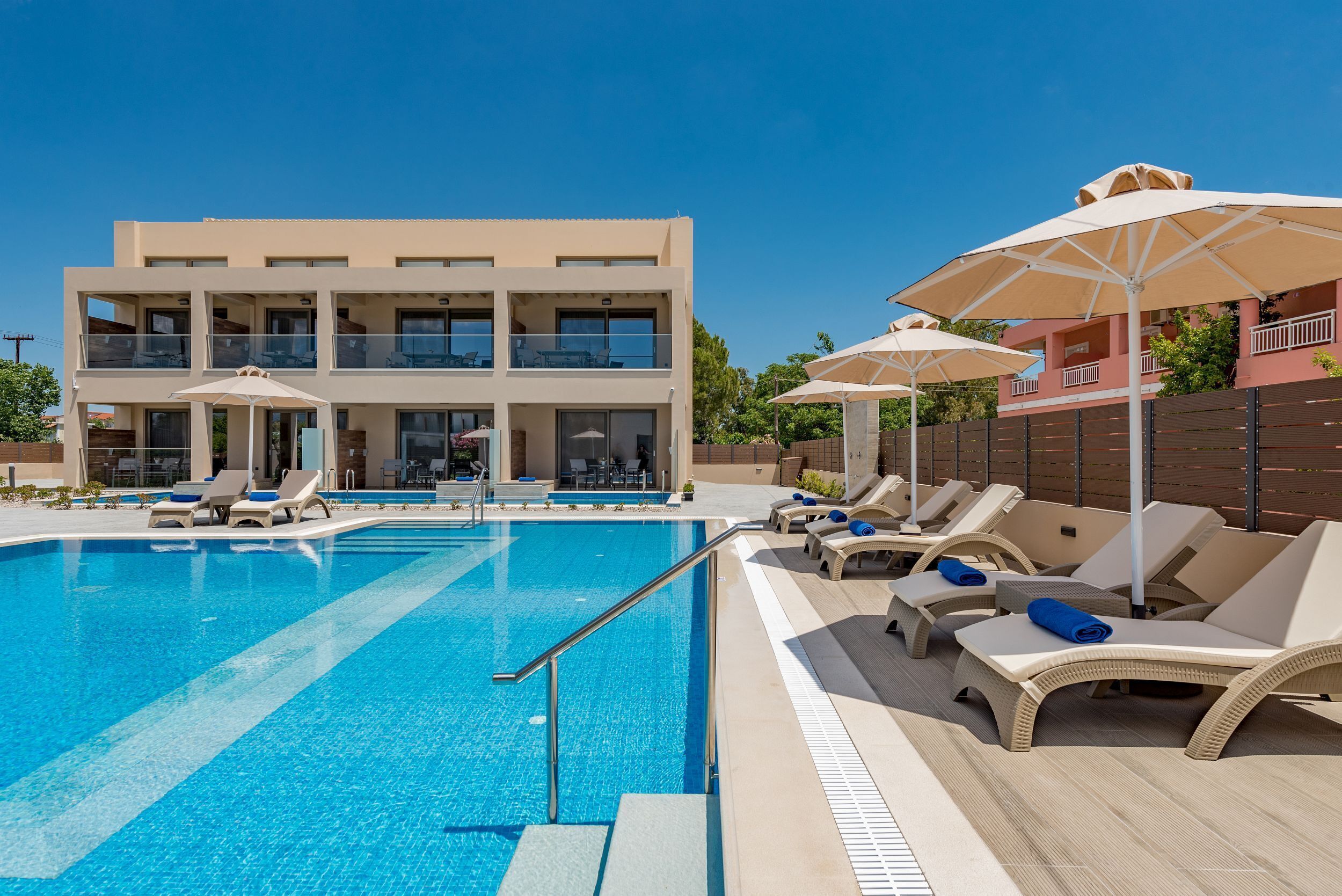 Une piscine avec des chaises longues et des parasols, un immeuble à plusieurs étages doté de balcons, et un ciel bleu azur.