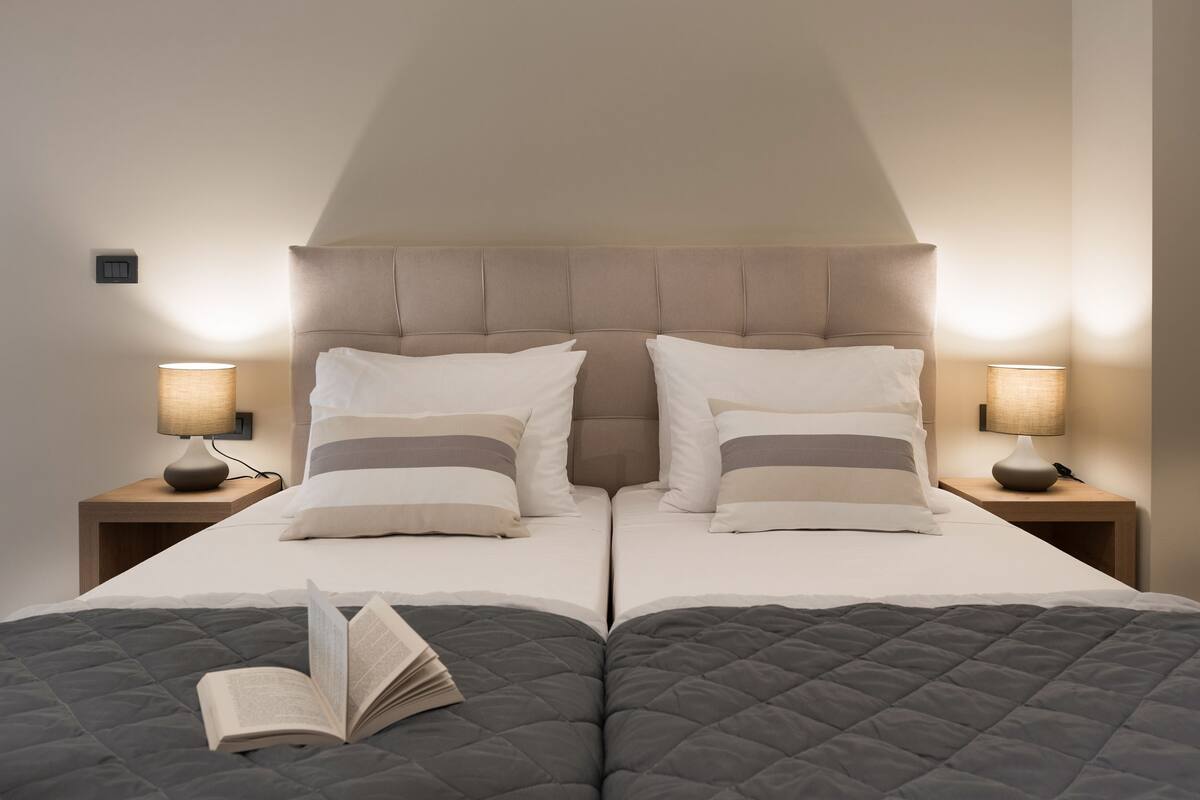 Un letto matrimoniale con copriletto grigio trapuntato, due cuscini bianchi e un libro sul letto. Due comodini con lampade sono posti ai lati del letto.