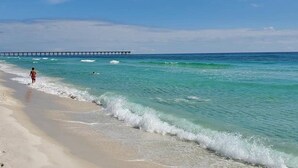 Een strand met helder turquoise water, een zandstrand en een pier op de achtergrond.