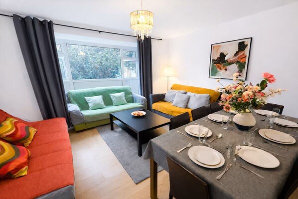 Ein modernes Wohnzimmer mit einem grünen Sofa, einem gelben Sofa, einem roten Sofa, einem Esstisch gedeckt mit Tellern und Gläsern sowie einem Chandelier.