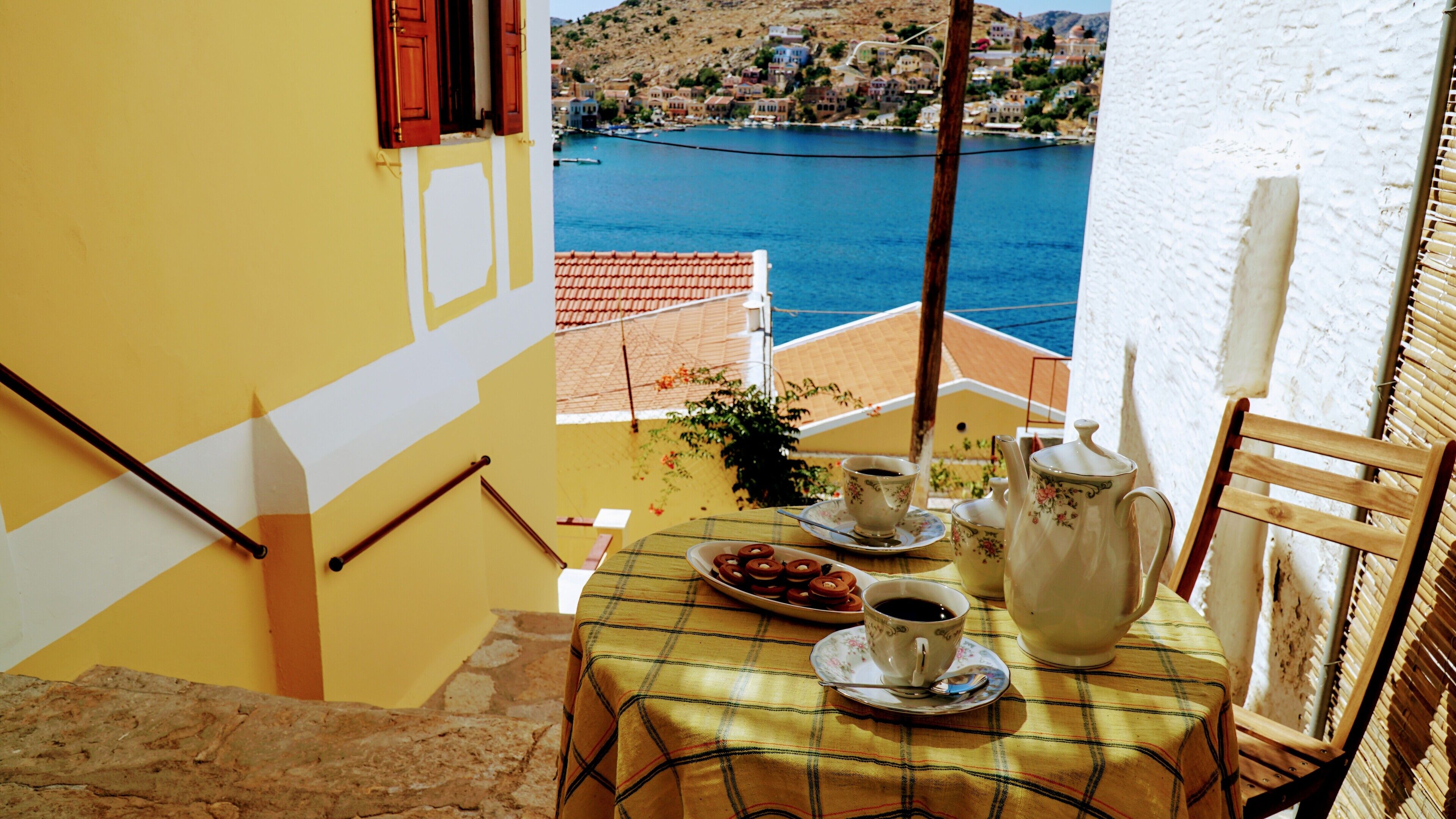 A balcony with a table set for breakfast, overlooking a coastal town.