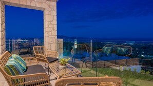 A terrace with wicker furniture, a glass railing, and a city view at dusk.