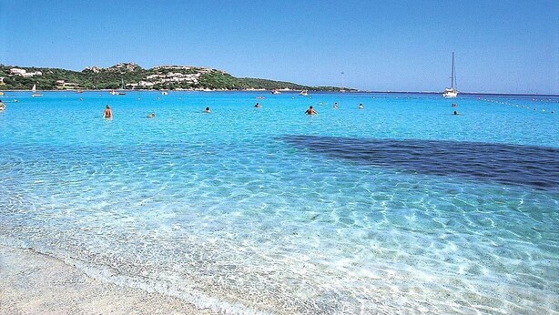 Une mer d’un bleu profond où l’on voit des gens nager et un voilier au loin.